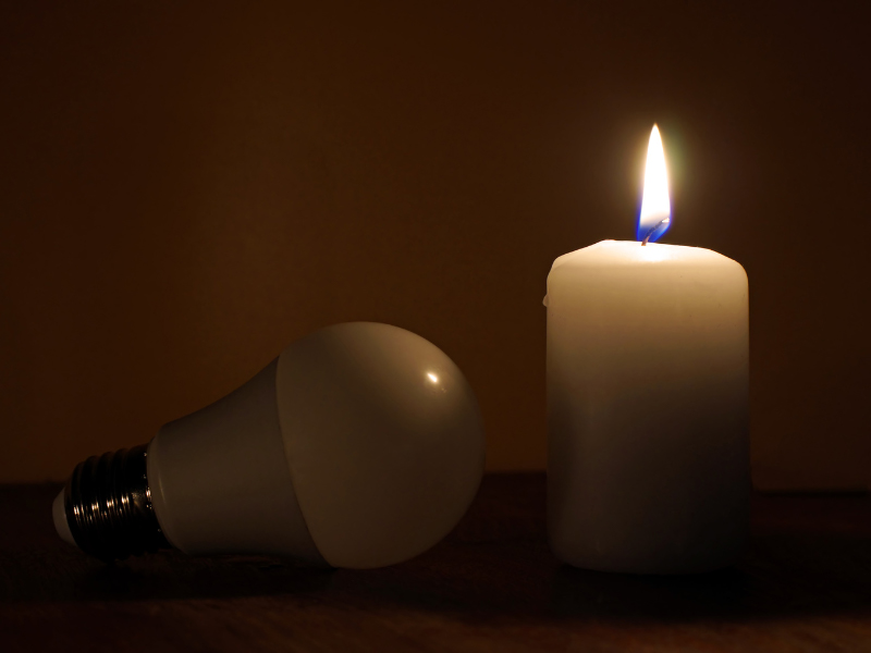 Eine brennende Kerze steht neben einer ausgeschalteten LED-Glühbirne in dunkler Umgebung – Symbolbild für Stromausfall.